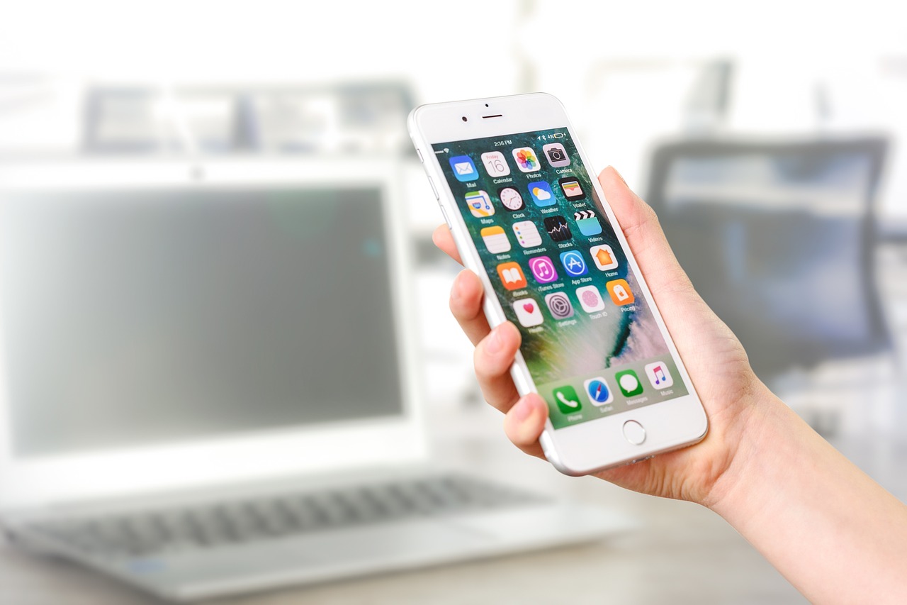 woman holding mobile phone displaying apps with laptop in background